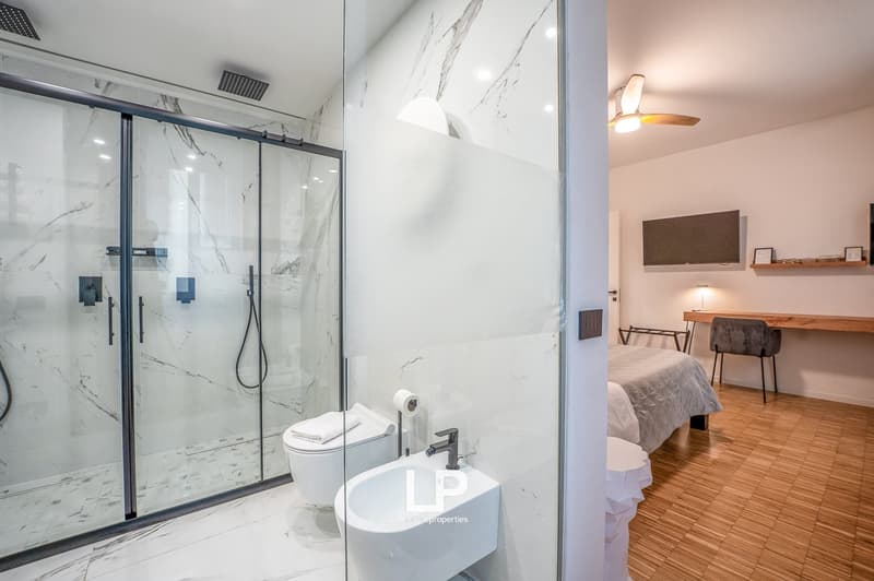 glass shower, white bidet, white sink, wooden floor, mounted TV, desk, chair