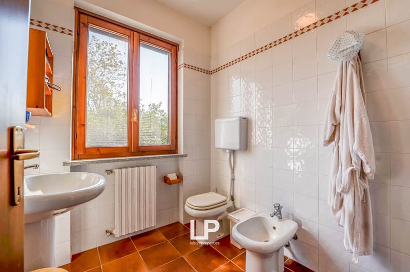 Bathroom with tiled walls, wooden door, a sink, a toilet, a bidet, a towel hanging on the wall, and a wooden window frame.