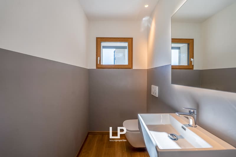 Bathroom with white walls, a white sink, a toilet, wooden floor, a mirror, two small windows with wooden frames
