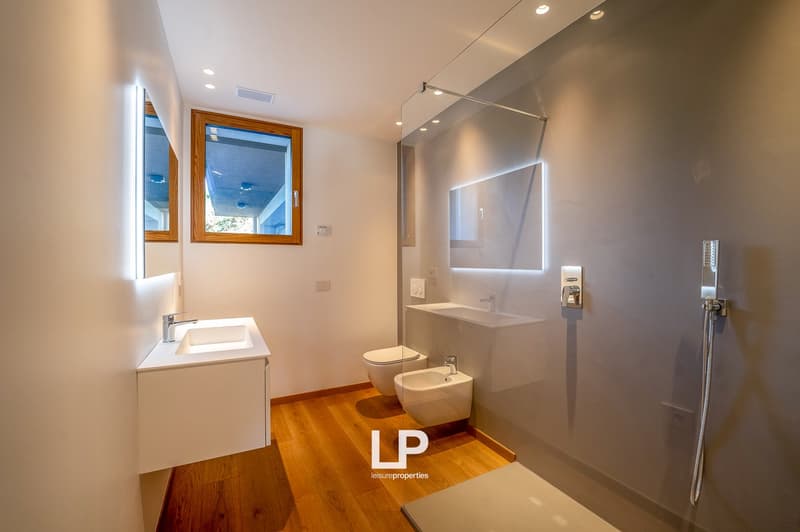 A small bathroom with wooden floors, white walls, a window, a vanity, a toilet, a bidet, and a shower with glass enclosure and showerhead