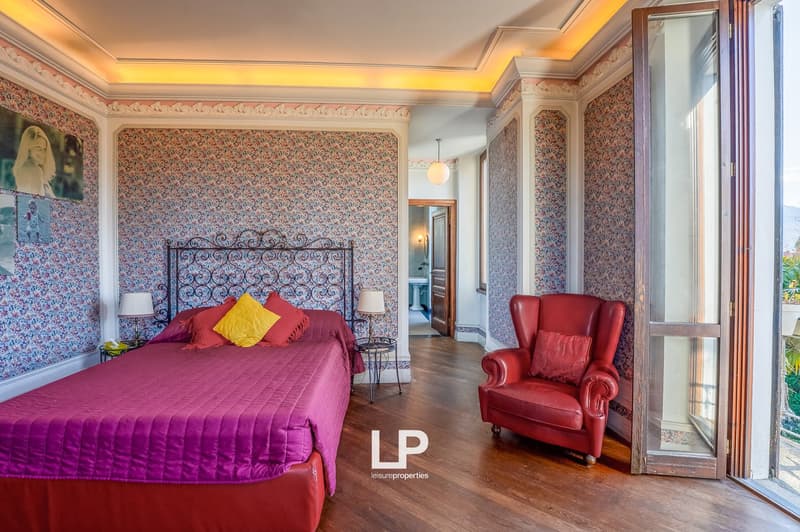 large bed, red and yellow pillows, bedside tables with lamps, red armchair, floral wallpaper, wooden floors, doorway to another room