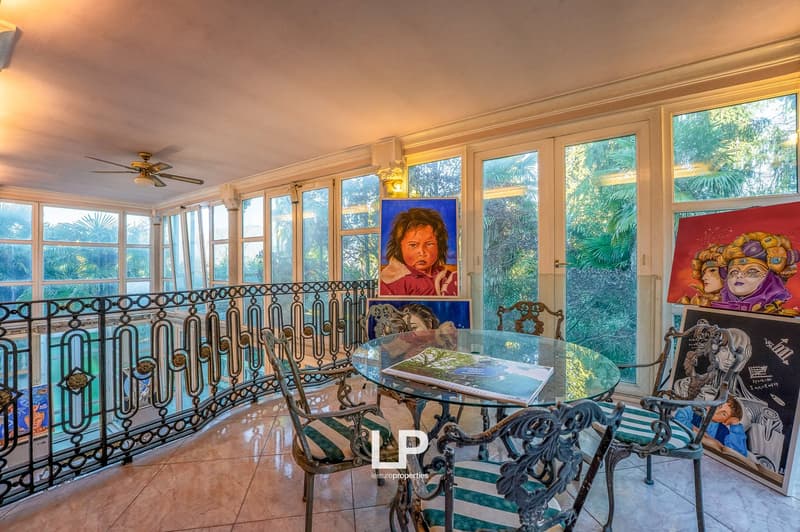 Decorated glass balcony with metal railings and large windows, paintings, wooden table, chairs, books on table