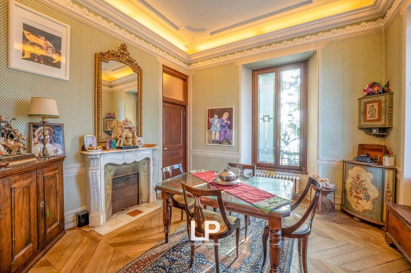 Dining room with wooden floor, fireplace, dining table, wooden chairs, decorative items, lamps, mirror, paintings, radiator, and window
