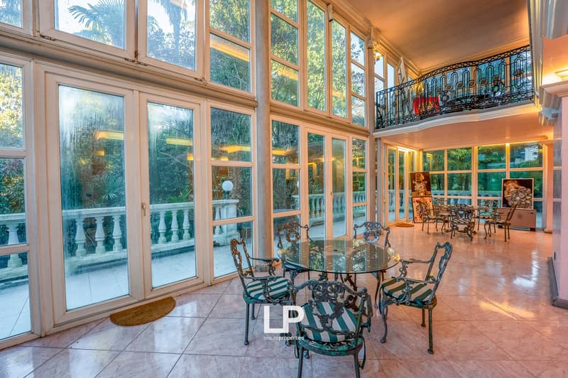 A room with large glass doors and windows, a glass table, chairs, and an open balcony with railings, offering a view of trees and the outdoors.