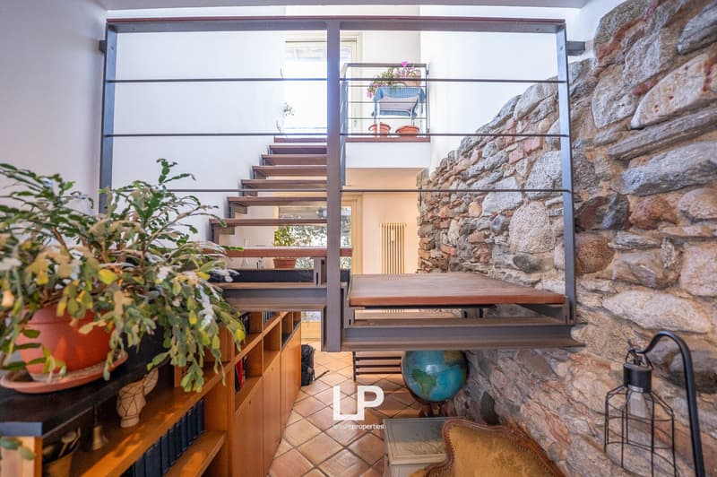 Raised ground floor, wooden staircase, stone wall, balcony, metal railings, indoor plants, books on a shelf, globe, vintage lamp
