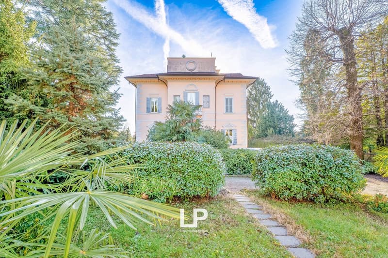 Large two-story house with trees and bushes, paved path to entrance, multiple windows, arched roof.
