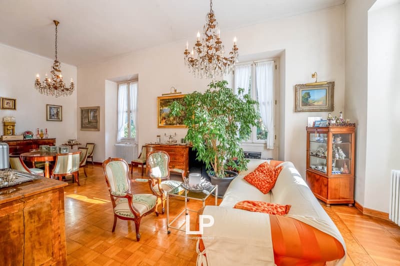 wooden floor, fireplace, large windows, chandeliers, paintings, sofa, chairs, table, cabinet, and a plant