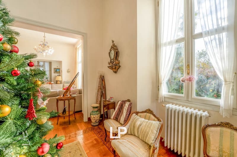 Living room: Christmas tree, wooden floor, radiator, windows, curtains, sofa, decorative items, lamp, dining table, clock on the wall, vase, armchairs