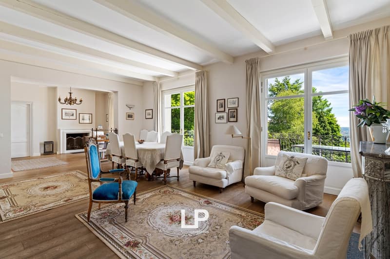 High ceilings, wooden floors, large white living chairs, chandelier, rug, fireplace, large windows with balcony, wooden table and chairs, desk with lamp, plants