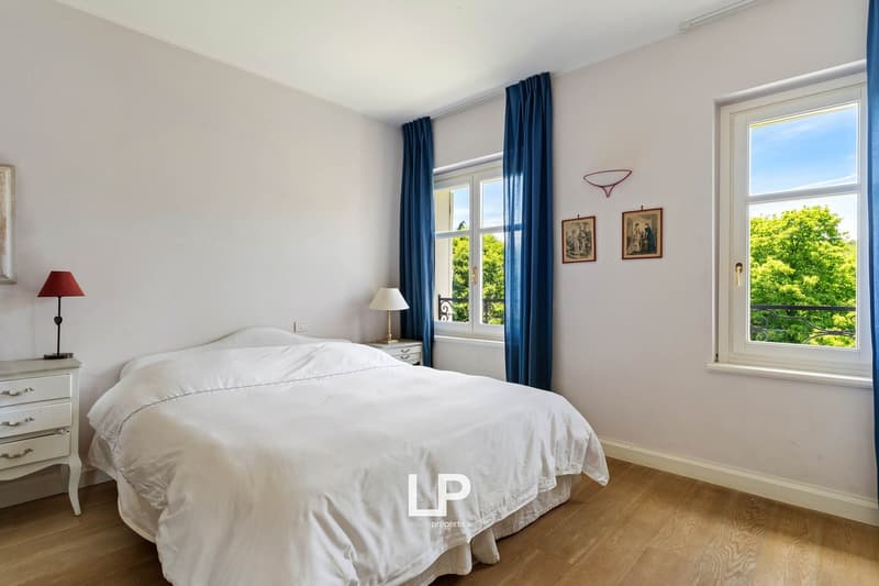 Double bed with white bedding, white bedside tables with lamps, wooden floor, white walls, two windows with blue curtains, balcony with black railings, outdoor view