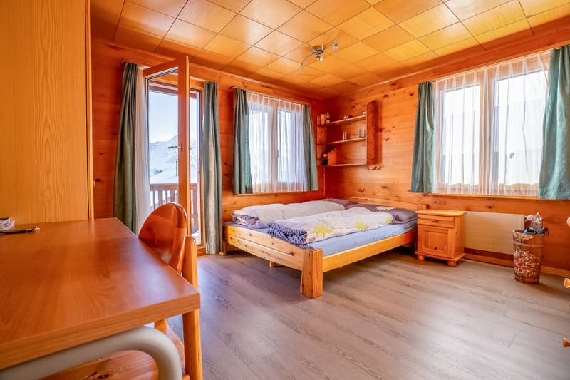 bedroom with a double bed, wooden walls, desk, wooden table, chair, two windows, balcony