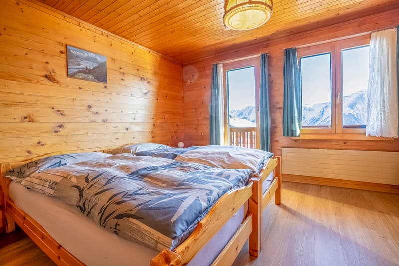 bedroom with wooden wall, two beds, curtains, windows with balcony view of mountains