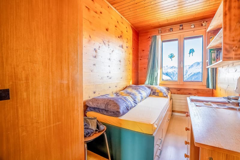 Bedroom with single bed, wooden walls, two windows with curtains, desk, wooden shelves, clothes hanging on a stool.