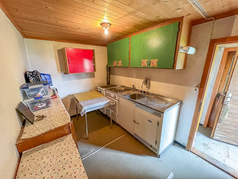 A small kitchen with wooden cabinets, a sink, stove, and refrigerator, as well as two tables with a tablecloth, a lamp, and a few items on top.