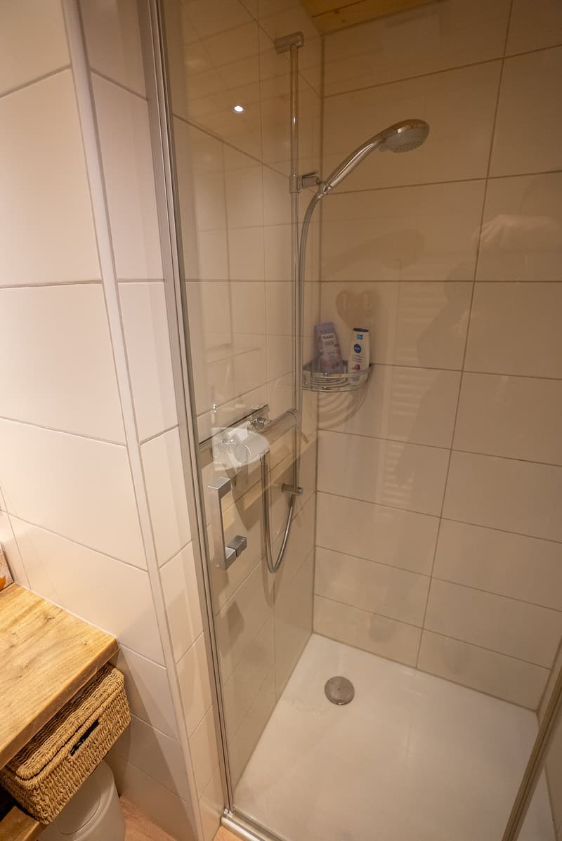 A shower stall with tiled walls, glass door, shower head, and drain, basket, white shelves
