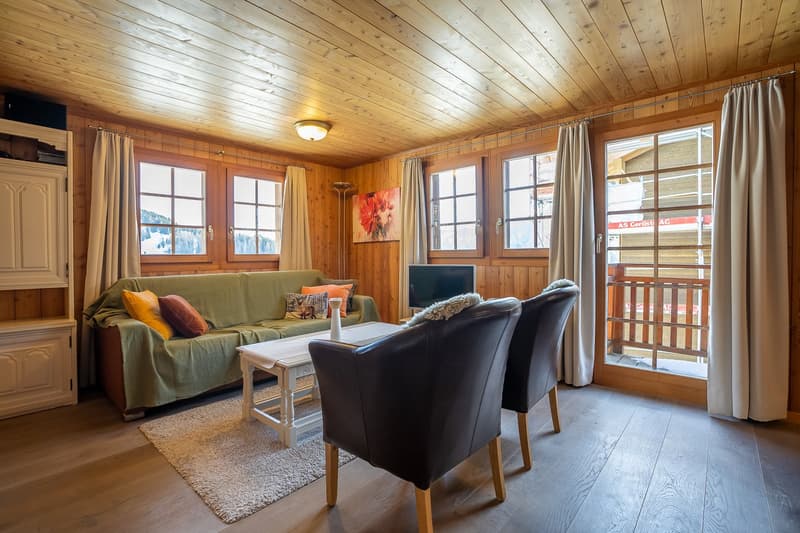 a living room with wooden floor, wooden ceiling, large windows, sofa, coffee table, rug, a tv mounted on the wall, balcony, and view of the outside