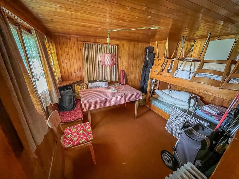 A room with wooden paneled walls, wooden floor, bunk beds, a desk, chair, and various items like pillows, storage boxes, and bags