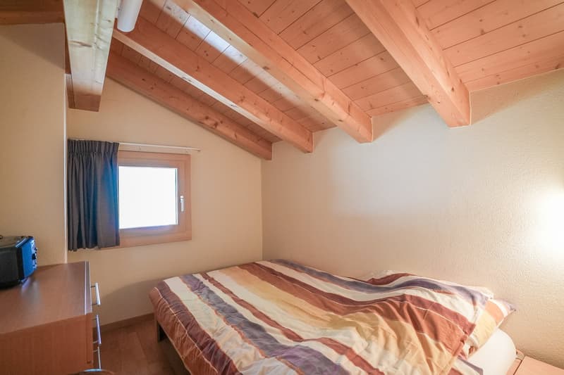 Small bedroom with wooden bed, bedside cabinet with drawers, window with curtains, exposed wooden beams, wooden flooring