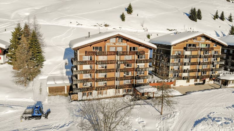 Hotel Valaisia, wooden exterior, snow on roof, several floors, balconies, set in snowy landscape.
