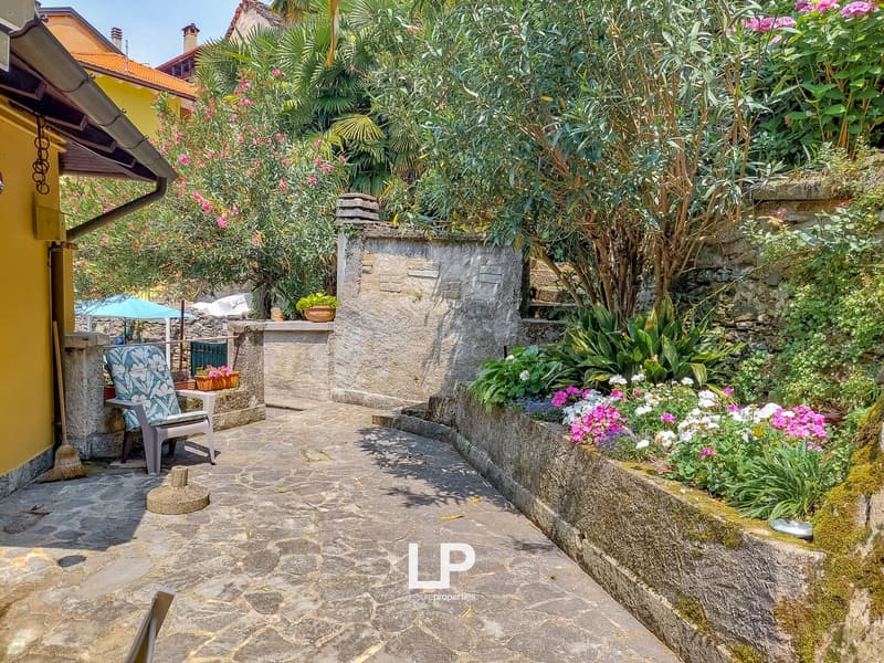Stone pathway, yellow house wall, potted plants, garden bed with flowers, greenery