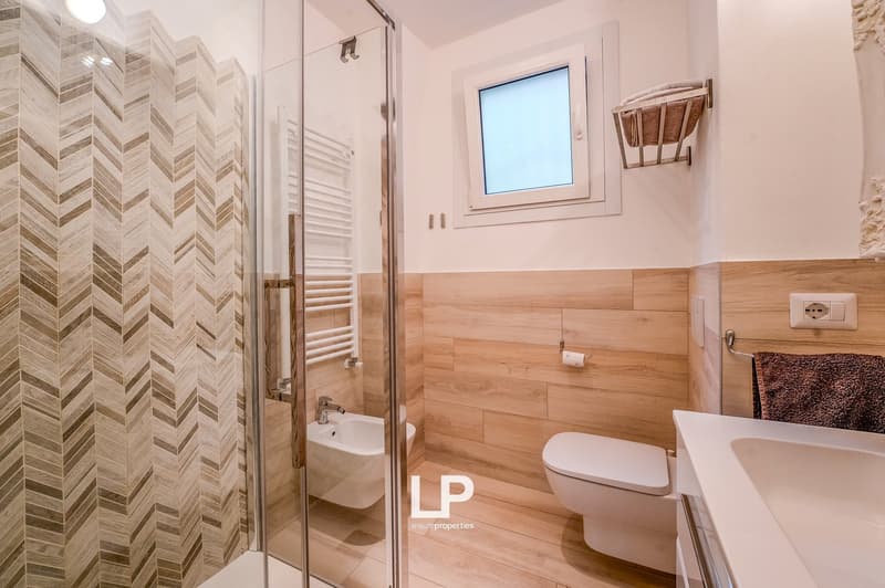 A modern bathroom with glass shower enclosure, white sink, a toilet, bidet, wooden walls and floors, towel rack, and window.