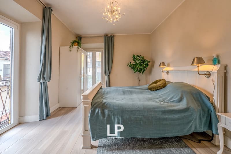 A bedroom with a bed, a wardrobe, and a window leading to a balcony. The bed is covered with a blue blanket, and there is a small pillow on it. The wall has two lamps and a plant. The floor has a wooden texture. The chandelier is hanging on the ceiling. T