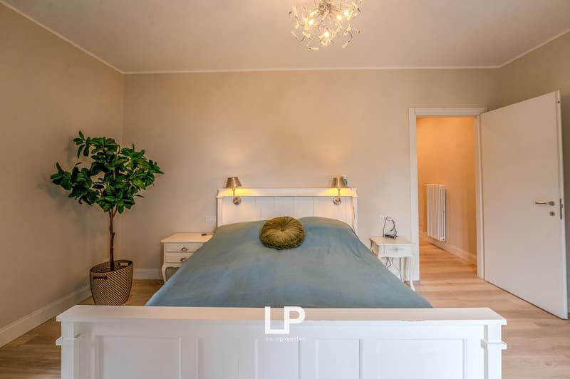 Large bed, two nightstands, lamp, radiator, plant, white walls, wooden floor