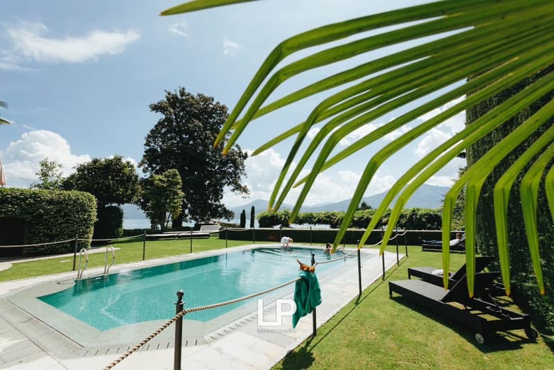 Outdoor swimming pool with green hedges, lawn, lounge chairs, trees, mountains, and a lake in the distance