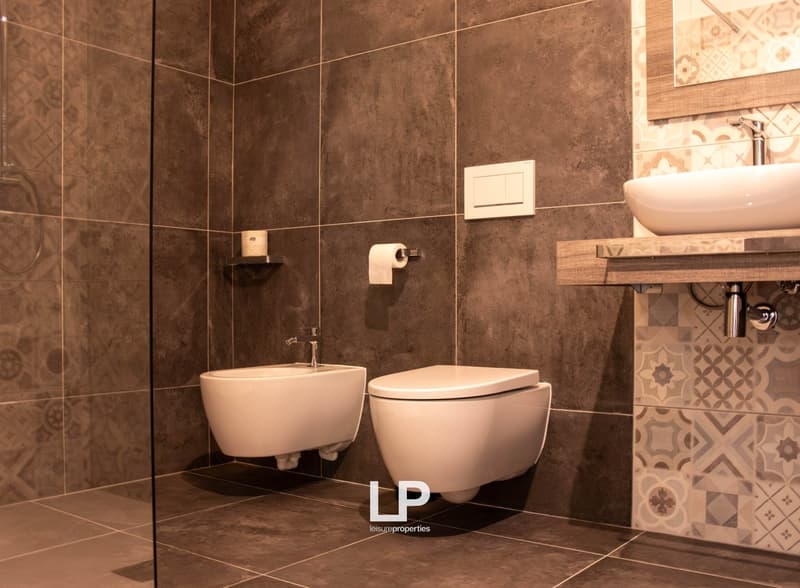 modern design, grey tiled walls, white fixtures, glass walk-in shower, bidet, toilet, sink, mirror, wooden shelf, and white roll of toilet paper