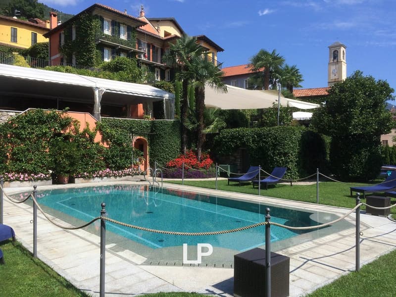 Swimming pool, green grass, several lounge chairs, surrounded by plants, large building with balconies