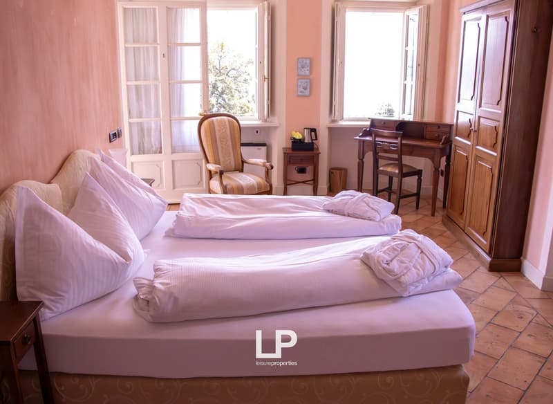 Two beds with white linen, wooden desk with chair, wooden wardrobe, two windows with curtains, wooden nightstands, pink walls, white pillow and blanket.