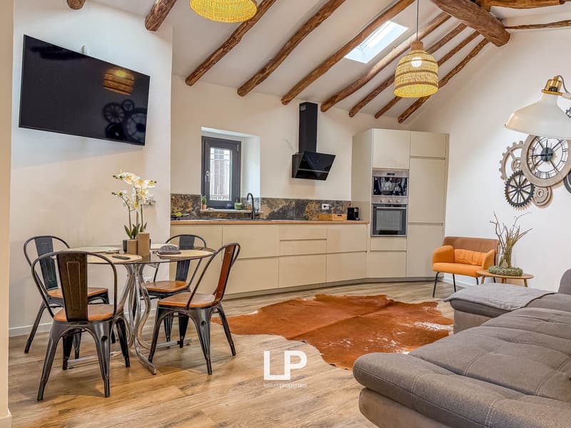 Open kitchen with dining area, living room with sofa and table, TV mounted on wall, wooden floor, chandeliers, skylight