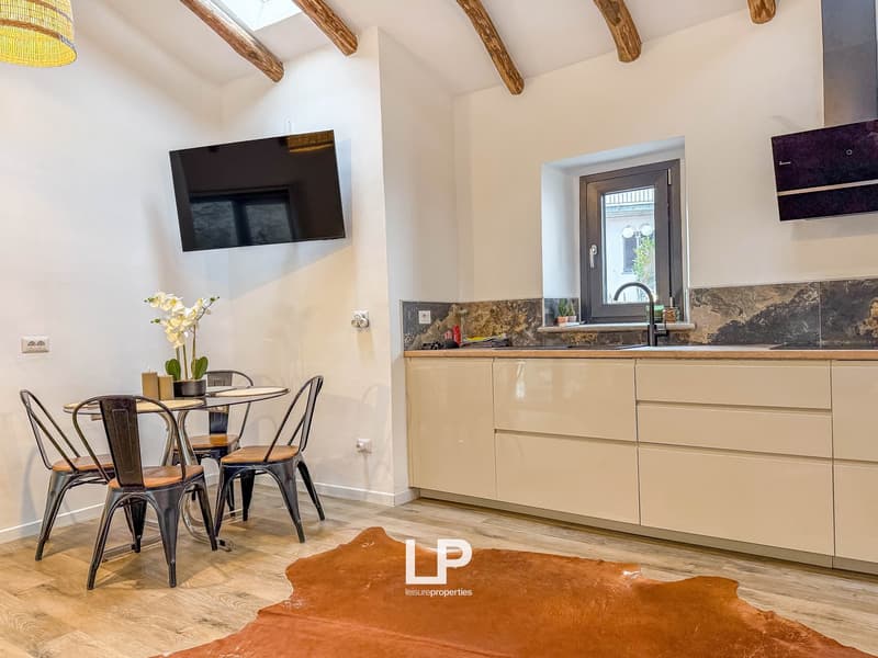Modern open kitchen with a dining area. It has a table and chairs, a TV mounted on the wall, a marble countertop, a sink, a faucet, and a small potted plant. It has wooden flooring, white walls, and a wooden ceiling with exposed beams.
