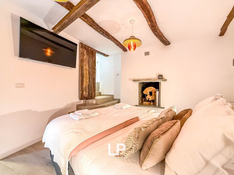 Bed with multiple pillows, fireplace in wall, wooden beams on ceiling, large tv on wall, wooden door leading to stairs