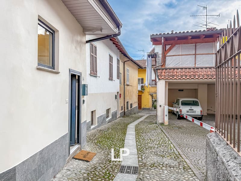 A narrow alley with cobblestone pavement, a small garage on the right side with a barrier gate, and houses on either side. One of the houses has a metal gate at the entrance, and a car is parked in front of it.