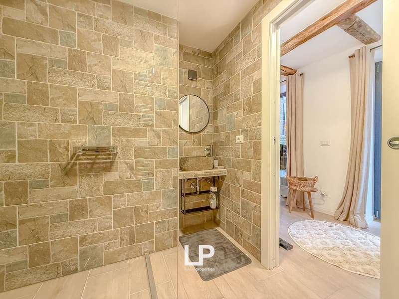 stone walls, walk-in shower, round mirror, stone sink, faucet, shelf with basket and bottle, rug, open door to another room