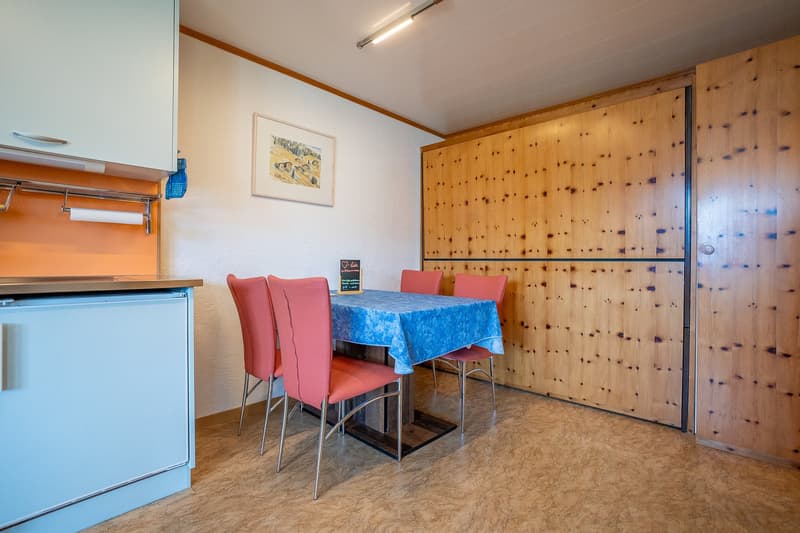 Dining area with wooden table and chairs, wooden cupboards, wooden wall paneling, small kitchen area with sink and refrigerator