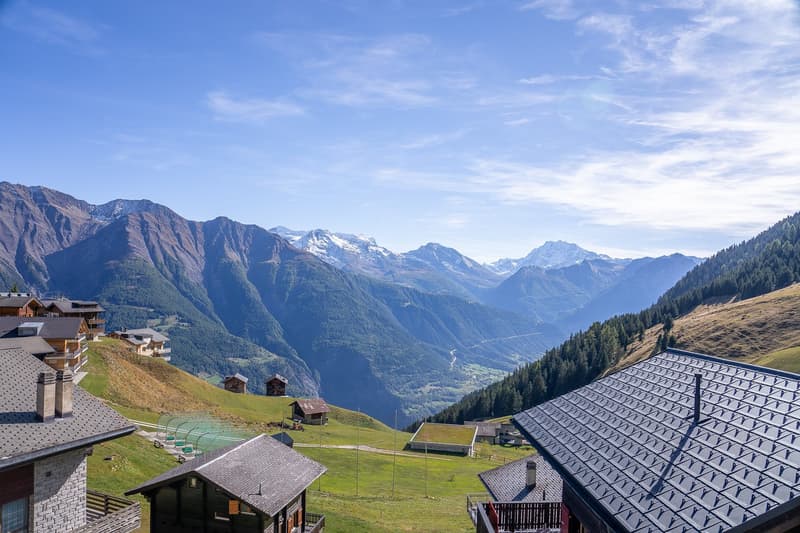 Overlooking a valley with mountains, alpine buildings, green grass, and a clear blue sky.