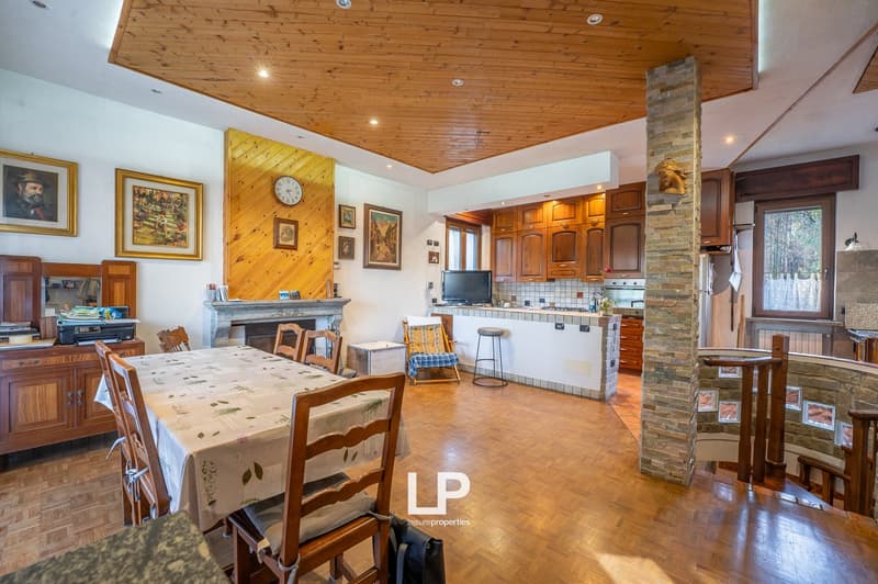 Dining area with wooden floors, dining table, chairs, fireplace, wall decorations, cabinets, kitchen island with stove, countertop, monitor, bar stool, staircase with railings, kitchen cabinets, windows, framed picture.