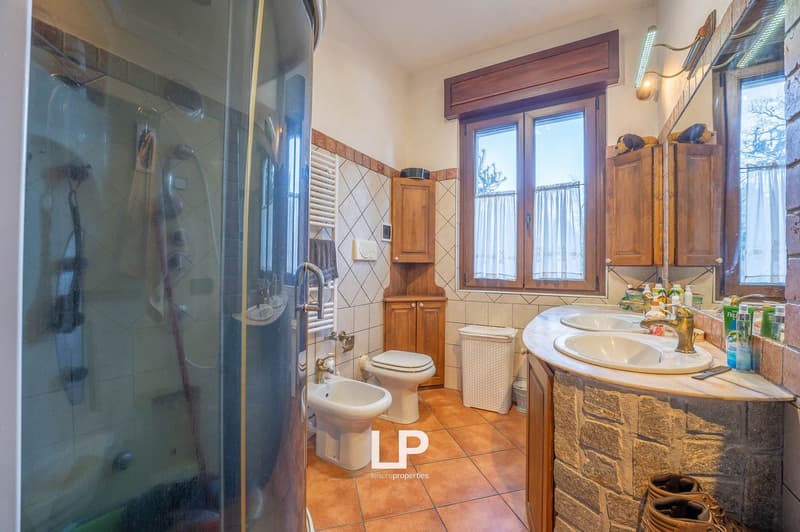 Modern bathroom with walk-in shower, 2 sinks, bidet, toilet, and wooden cabinets. Tiled floor and natural lighting from windows.