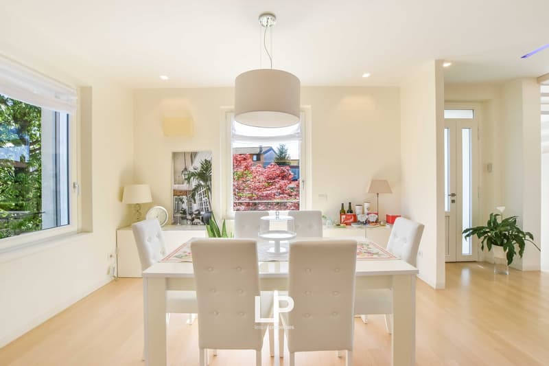 Bright and spacious dining room with large windows overlooking a scenic outdoor view. The room features a white dining table with four matching chairs, a pendant light fixture, and decorative elements such as plants and artwork on the walls.
