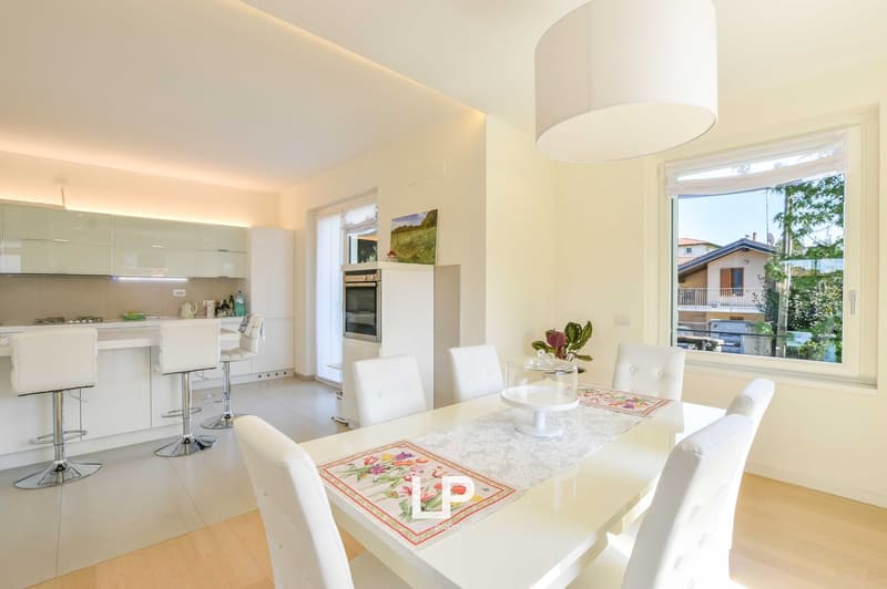 Open-concept kitchen and dining area with white cabinets, stainless steel appliances including a dishwasher, large windows providing natural light, and a dining table with white chairs.