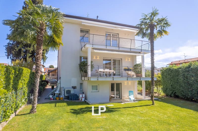 Two-story modern house with a balcony, surrounded by palm trees and a well-maintained lawn. The house has a clean, white exterior with large windows and a covered patio area on the ground floor.