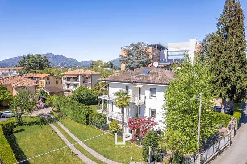 The image shows a residential area with multi-story apartment buildings, surrounded by lush greenery and trees. The buildings have balconies and terraces, and there appears to be a view of mountains or hills in the background.