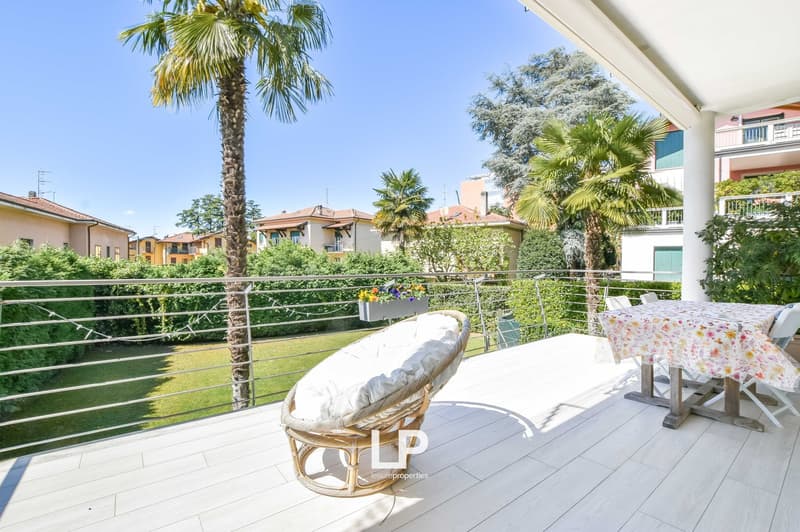 Spacious balcony with rattan furniture, overlooking a lush garden with palm trees. The apartment building has a modern, Mediterranean-style architecture with white walls and colorful accents.