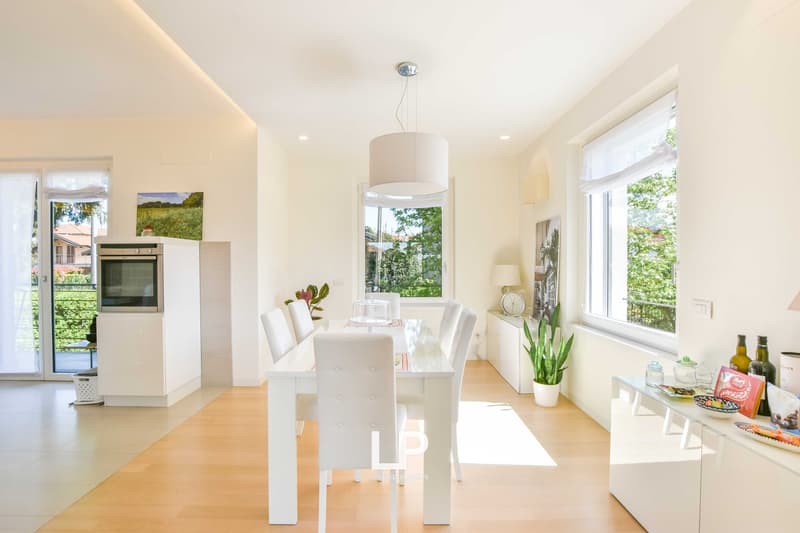 Bright and airy dining room with large windows overlooking a garden, white dining table and chairs, pendant light fixture, kitchen appliances visible in the background