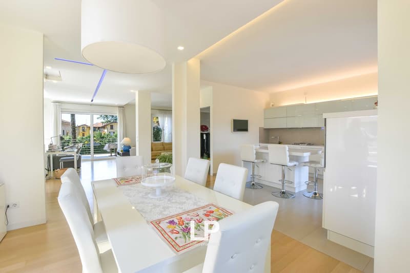 Spacious open-plan living area with dining table, chairs, and kitchen visible. Large windows provide natural light and access to a balcony or terrace. The room has a modern, minimalist design with white walls and floors.