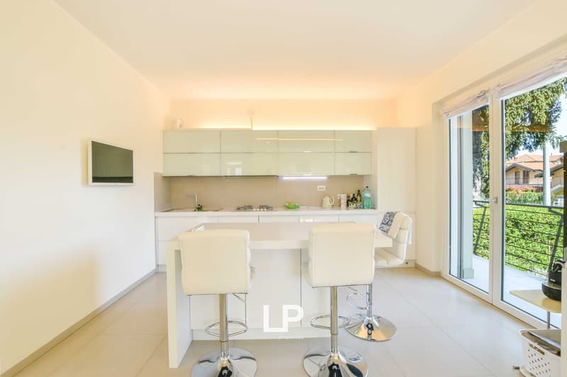 Bright and modern kitchen with white cabinets, stainless steel appliances, and a breakfast bar with two white stools. Large windows provide natural light and access to a balcony or terrace area.