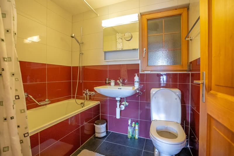 Bathroom with bathtub, sink, toilet, red tiled walls, white tiles on floor, overhead shower, mirror, window, toiletries