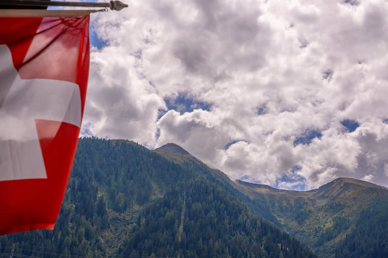 A red and white Swiss flag with a mountain range covered in trees and a cloudy sky in the background